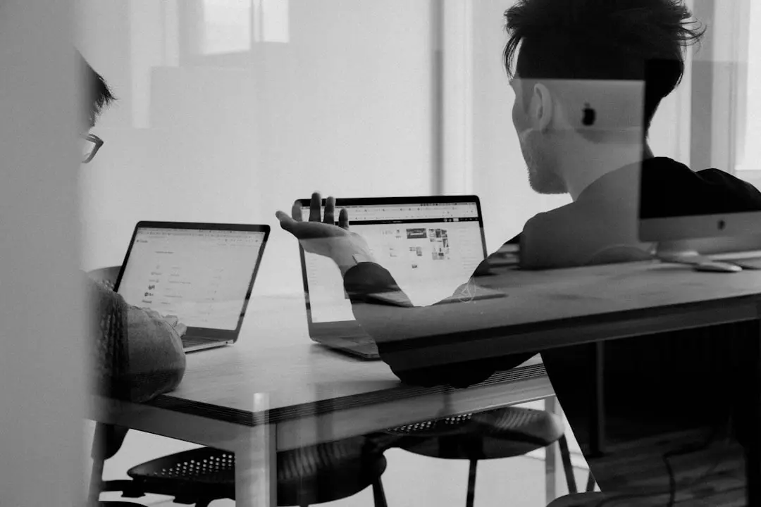 About Black and white photo of two professionals discussing work while using laptops in a modern office, seen through a glass reflection.
