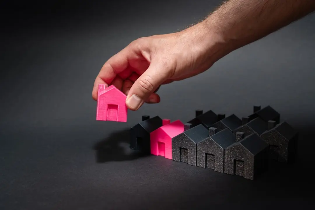 About A hand selecting a bright pink miniature house from a group of black houses, symbolizing real estate investment, housing market choices, or property differentiation.